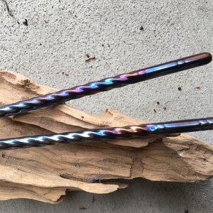 May include: Two twisted metal chopsticks with a blue, purple, and gold iridescent finish. The chopsticks are lying on a piece of driftwood.