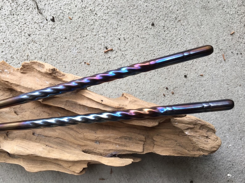 May include: Two twisted metal chopsticks with a blue, purple, and gold iridescent finish. The chopsticks are lying on a piece of driftwood.