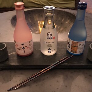 May include: Three small bottles of sake on a black slate serving tray with two chopsticks. The bottles are labeled "Draft Sake", "Yume No Yori", and "白鶴".