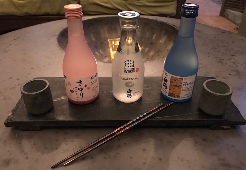 May include: Three small bottles of sake on a black slate serving tray with two chopsticks. The bottles are labeled "Draft Sake", "Yume No Yori", and "白鶴".
