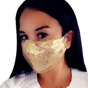 May include: A gold sequined face mask with a white elastic band. The mask is worn by a person with dark hair.