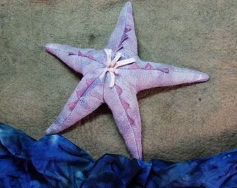 Puede incluir: Una estrella de mar de peluche de color púrpura con un centro rosa y detalles blancos. La estrella de mar está tumbada sobre una superficie marrón con una tela azul en primer plano.