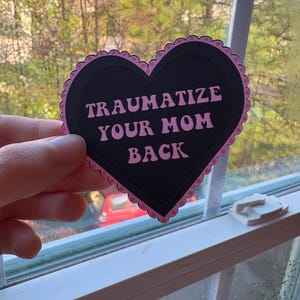 Puede incluir: Una pegatina negra en forma de corazón con bordes festoneados rosas y la frase "TRAUMATIZE YOUR MOM BACK" en texto rosa. La pegatina se sostiene frente a una ventana con un fondo exterior borroso.