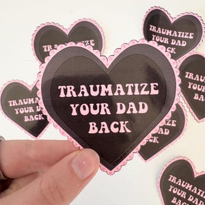 Puede incluir: Un conjunto de pegatinas en forma de corazón con fondo negro y bordes festoneados rosas. Las pegatinas presentan el texto "TRAUMATIZE YOUR DAD BACK" en rosa. Se ven otras pegatinas en el fondo.