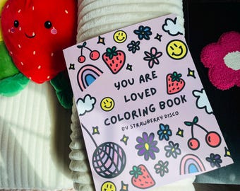 Puede incluir: Un libro para colorear rosa titulado "You Are Loved Coloring Book" de Strawberry Disco, con ilustraciones de fresas, arcoíris, flores y caras sonrientes. También se ve una fresa de peluche y una flor rosa.