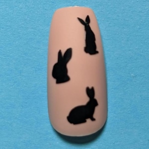 May include: A single artificial nail with a light peach color, adorned with three black rabbit silhouettes. The nail is set against a solid blue background, creating a contrasting visual. The design is simple and elegant.