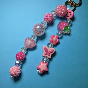 Op de afbeelding: Een roze kralen sleutelhanger met een goudkleurige sluiting. De sleutelhanger heeft verschillende roze kralen, waaronder getextureerde bollen, heldere kristallen kralen en bedels in de vorm van een hart, vlinder, ster, bloem, aardbei en een smiley.
