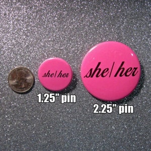 K&ouml;nnte beinhalten: Zwei rosa Anstecknadeln mit dem Text "she/her" in Schwarz. Die kleinere Nadel hat einen Durchmesser von 1,25 Zoll und die gr&ouml;&szlig;ere einen Durchmesser von 2,25 Zoll.