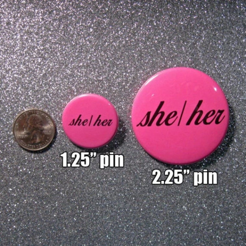 K&ouml;nnte beinhalten: Zwei rosa Anstecknadeln mit dem Text "she/her" in Schwarz. Die kleinere Nadel hat einen Durchmesser von 1,25 Zoll und die gr&ouml;&szlig;ere einen Durchmesser von 2,25 Zoll.