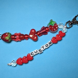 May include: Two beaded keychains on a blue background. One keychain features a strawberry charm, red beads, and a lobster clasp. The other keychain has red heart beads and the word "CUSTOM" spelled out in white letter beads.