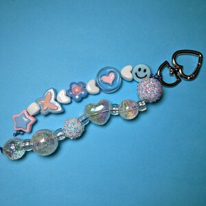 May include: A beaded keychain with charms including a star, butterfly, flower, heart, and smiley face. The keychain has a silver heart-shaped clasp and is set against a blue background. The beads are in pastel colours.