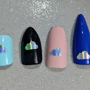 May include: Four press-on nails in different shapes and colors, each featuring a small holographic cloud decal. The set includes a light blue square nail, a black almond nail, a matte pink coffin nail, and a long blue stiletto nail.