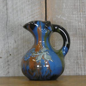 May include: A small blue and brown ceramic pitcher with a leafy design. The pitcher has a black handle and a slightly flared lip.