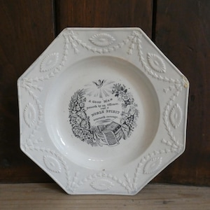 May include: An octagonal white ceramic plate featuring a central black transfer print of a floral wreath, a dove, and the text: A GOOD MAN passeth by an Offence. and a NOBLE SPIRIT scorneth revenge. The rim has embossed floral and leaf garlands.