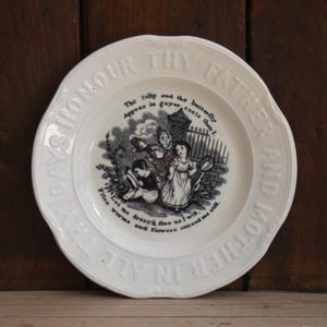 May include: A white ceramic plate with embossed text around the rim reading HONOUR THY FATHER AND MOTHER IN ALL THY DAYS. The centre features a black and white illustration of two children in a garden with text about tulips and butterflies.