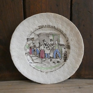 May include: A white ceramic plate with a textured, dimpled border featuring a central illustration titled THE BOTTLE. The scene depicts figures in period clothing with text reading PLATE VII-THE HUSBAND IN A STATE OF FURIOUS DRUNKENNESS and KILLS HIS WIFE WITH THE INSTRUMENT OF ALL THEIR MISERY.