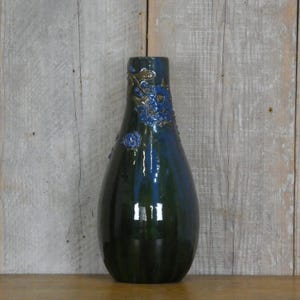 Könnte beinhalten: Eine hohe, dunkelgrüne Keramikvase mit glänzendem Finish. Die Vase hat blaue Blumenverzierungen am oberen Rand. Die Vase hat einen abgerundeten Körper und einen schmalen Hals, ideal für Blumen.