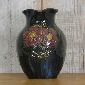 May include: A dark blue ceramic vase with a floral design. The vase features three red flowers with yellow centres and green leaves.