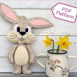 Op de afbeelding: Een PDF-patroon voor het naaien van een vilten konijnenknuffel. Het konijn is grijs met roze oren en een witte buik. Het heeft zwarte ogen en een zwarte neus. Het konijn staat op een wit houten oppervlak. Er zijn gele bloemen in een witte kan met een bloemenpatroon.