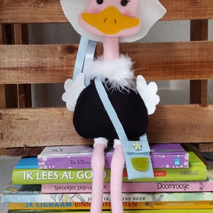 Könnte beinhalten: Eine rosafarbene Straußenpuppe aus Filz, die einen weißen Hut mit einer blauen Blume, ein schwarzes Kleid und eine blaue und grüne Tasche trägt. Die Puppe sitzt auf einem Stapel bunter Bücher.