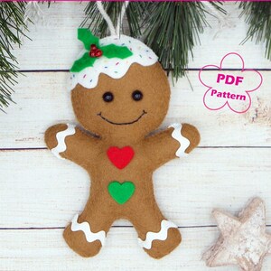 May include: A felt gingerbread man ornament with a white icing top, red and green felt hearts, and a holly sprig on top. The ornament is hanging from a white string. The text "PDF Pattern" is visible in the image.