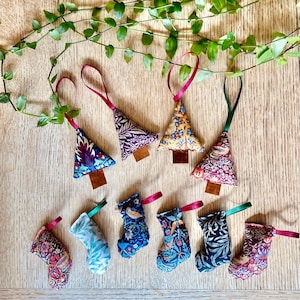 May include: A collection of four fabric Christmas tree ornaments and six fabric stocking ornaments, each featuring intricate floral and bird patterns in various colours, with attached ribbon loops for hanging and small brown rectangular bases.