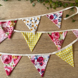 May include: Three fabric bunting banners with triangular flags. The flags display floral patterns in pink, red, and white, and yellow gingham. The banners are strung on a cream ribbon, ideal for celebrations.