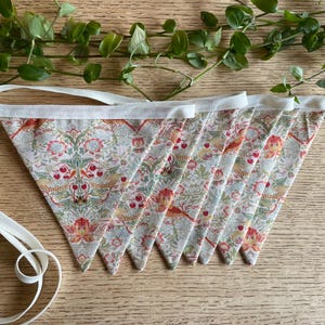May include: A decorative bunting featuring triangular fabric flags with a floral pattern in red, green, and cream. The flags are strung together with a white ribbon. The bunting is displayed on a wooden surface.