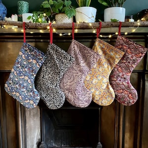 May include: Five festive Christmas stockings with patterned designs, suspended from a wooden branch. The stockings display floral and botanical patterns in blue, black, purple, yellow, and red. Red ribbons hang the stockings above a fireplace.
