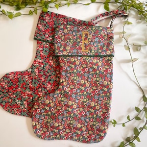 May include: Two Christmas stockings made of fabric with a dense floral pattern in shades of red, blue, yellow, and green. The front stocking features a dark green cuff with the letter E embroidered in gold thread and a hanging loop at the top.