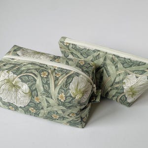 May include: Two fabric cosmetic bags with a floral pattern. The bags are rectangular with a zip closure. The fabric features a green background with white and yellow flower and leaf designs.