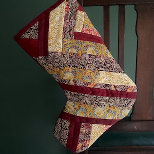 May include: A patchwork Christmas stocking in burgundy, gold, and cream. The stocking is decorated with floral and geometric patterns. It is hanging on a wooden chair, showcasing its festive design. The stocking is a handmade item.