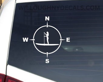 May include: White vinyl sticker of a compass with a woman paddleboarding in the center. The compass points are labeled N, S, E, and W.