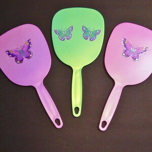 Pode incluir: Três espelhos de mão com desenhos de borboletas. Os espelhos são em tons de rosa, roxo e verde. Cada espelho tem um desenho de borboleta com asas roxas e detalhes amarelos.