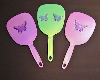 Pode incluir: Três espelhos de mão com desenhos de borboletas. Os espelhos são em tons de rosa, roxo e verde. Cada espelho tem um desenho de borboleta com asas roxas e detalhes amarelos.