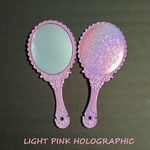 Pode incluir: Dois espelhos de mão ornamentados com um acabamento holográfico rosa claro. Os espelhos têm um design floral e uma borda festoneada.