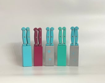 Pode incluir: Seis pequenas facas decorativas com cabos pintados. Os cabos são pintados em várias cores, incluindo azul, turquesa e bordô. As facas estão dispostas em uma fileira sobre uma superfície branca.