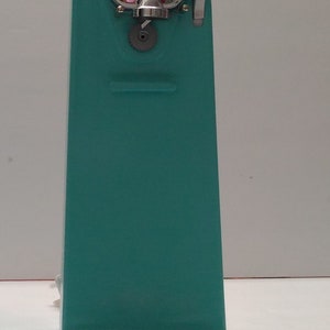 Pode incluir: Um abridor de latas manual azul turquesa com uma pega cromada e uma engrenagem na frente.