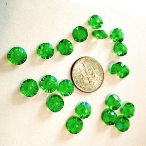 May include: A collection of 18 green faceted glass beads, each with a hole in the center. The beads are arranged in a circle around a US dime.
