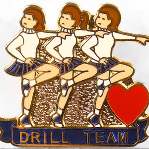 May include: Gold-colored pin with three female figures in blue and white skirts, performing a drill team routine. The figures are standing on a gold background with a red heart and a blue banner that reads "DRILL TEAM".