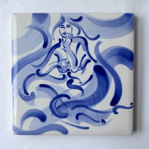 Puede incluir: Azulejo cerámico cuadrado con una ilustración azul y blanca de una mujer con cabello ondeante. La obra de arte representa una figura con una mano en la barbilla, rodeada de olas y líneas azules estilizadas. El azulejo tiene un acabado brillante.