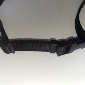 May include: Close-up of a black adjustable strap with a plastic buckle. The strap is made of a woven fabric.