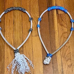 Könnte beinhalten: Zwei Seilhalfter mit dekorativen Akzenten. Ein Halfter hat silberfarbene Akzente und eine weiße Quaste, das andere blaue Akzente. Beide sind aus weißem Seil gefertigt und liegen auf einer Holzoberfläche.