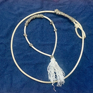 Könnte beinhalten: Ein weißes Seil mit dekorativen silbernen und grauen Akzenten und einer weißen Quaste. Das Seil ist geschlungen und auf einer blauen Oberfläche angeordnet. Das Seil wird wahrscheinlich für den Reitsport oder Western-Themen verwendet.