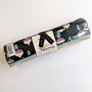 Może przedstawiać: Zwinięty pakiet tkaniny owinięty w przezroczysty plastik. Tkanina ma ciemne tło z powtarzającym się wzorem różowych i białych kształtów cebul i niebieskich wzorów przypominających rośliny. Etykieta głosi "Petunia by Kimberly Knight."