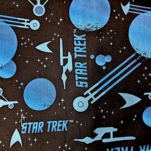 Könnte beinhalten: Ein schwarzer Stoff mit einem sich wiederholenden Muster aus blauen Sternen, Planeten und Raumschiffen. Der Text "STAR TREK" erscheint zweimal auf dem Stoff.