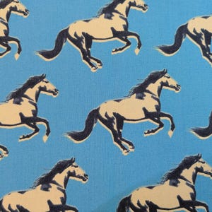 Puede incluir: Patrón repetido de caballos corriendo en negro y beige sobre un fondo azul claro. Los caballos están estilizados con crines y colas oscuras, creando un diseño dinámico y artístico.