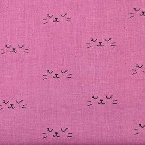 May include: A vibrant magenta fabric featuring a repeating pattern of small, black, hand-drawn cat faces. Each cat face consists of two closed eyes, a tiny nose, and three whiskers on each side, evenly spaced across the textured textile surface.
