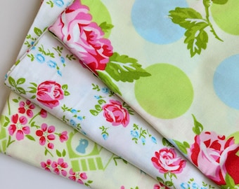 May include: Three fabric swatches with floral patterns. The top swatch has a white background with green leaves, pink roses, and blue polka dots. The middle swatch has a white background with pink roses and green leaves. The bottom swatch has a yellow background with pink flowers and green leaves.