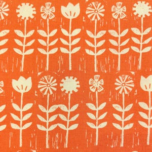 以下が含まれることがあります： 鮮やかなオレンジ色の生地に、クリーム色の花と茎がブロックプリント風のパターンで繰り返されています。様々な花びらの形をした様式化された花が列をなして並んでおり、手押しスタンプのような質感のあるデザインが特徴です。